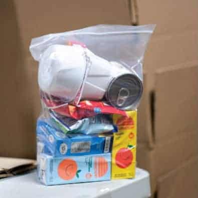 A clear plastic bag filled with empty food and drink containers, including juice boxes, a can, and disposable cups. Cardboard boxes are blurred in the background.