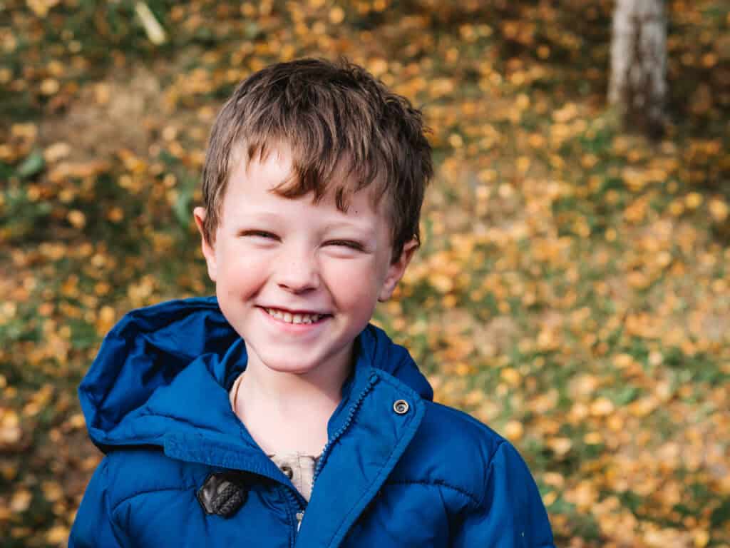 A young boy with brown hair, wearing a bright blue jacket, smiles outdoors. The background shows autumn leaves scattered on the ground and some greenery.