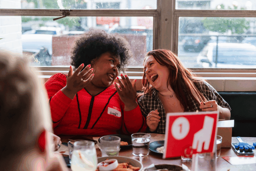 Two women sit together at a table in a restaurant, laughing and animatedly talking. Food and drinks are on the table, and a red table sign with the number 1 is visible in the foreground.