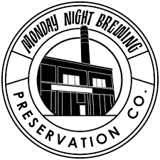 Black and white circular logo for Monday Night Brewing Preservation Co., featuring a stylized factory building with a tall smokestack in the center. The company name is written around the building.