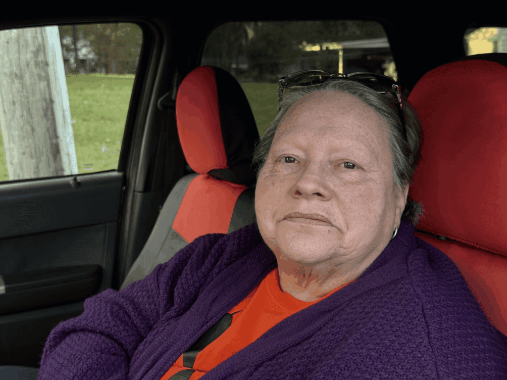 An older woman with light skin, short hair, and glasses resting on her head sits in a car with red and black seat covers, wearing a purple sweater and orange shirt, looking at the camera with a neutral expression.