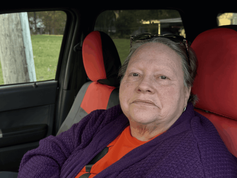 An older woman with light skin, short hair, and glasses resting on her head sits in a car with red and black seat covers, wearing a purple sweater and orange shirt, looking at the camera with a neutral expression.