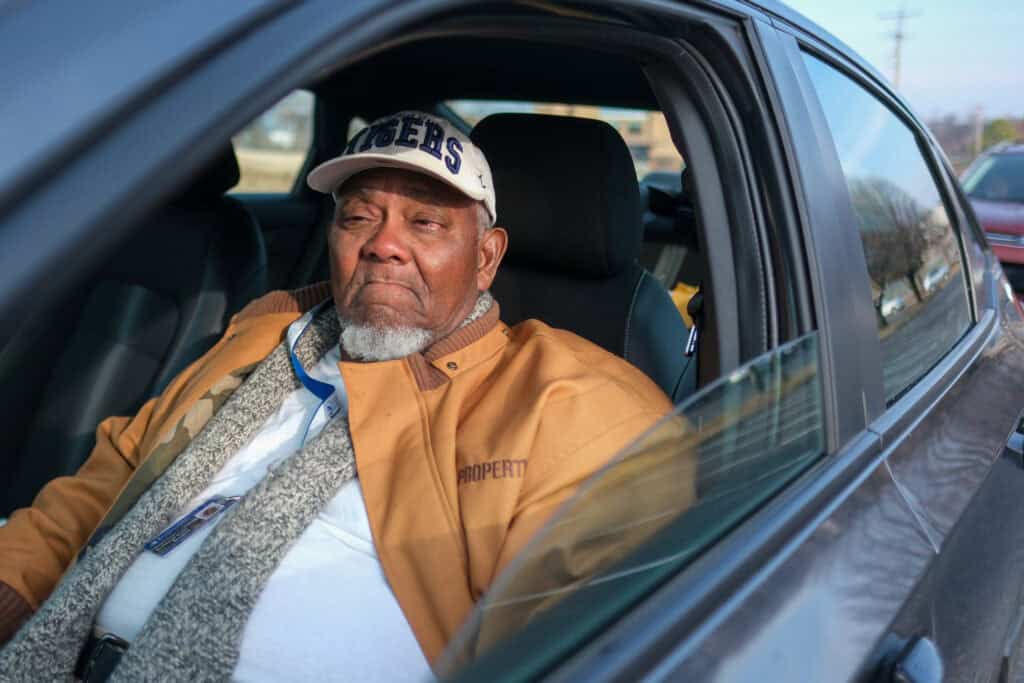 An elderly man wearing a tan jacket and a white cap sits in the drivers seat of a car, looking out the window with a thoughtful expression. Sunlight illuminates his face and part of the car interior.