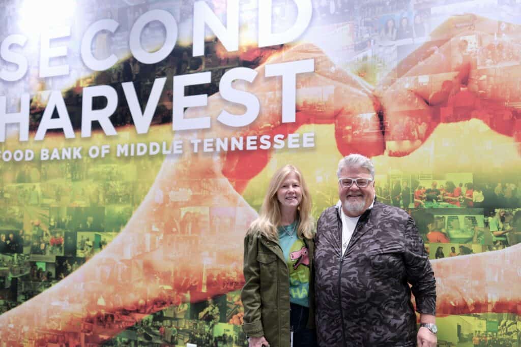A smiling woman and man stand in front of a colorful mural that reads Second Harvest Food Bank of Middle Tennessee. The background features large hands forming a heart shape and various community images.