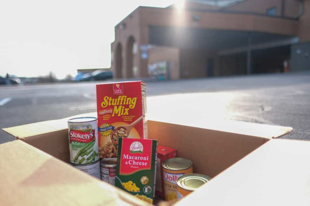 A cardboard box filled with canned food, stuffing mix, and macaroni & cheese sits in a parking lot outside a building on a sunny day.