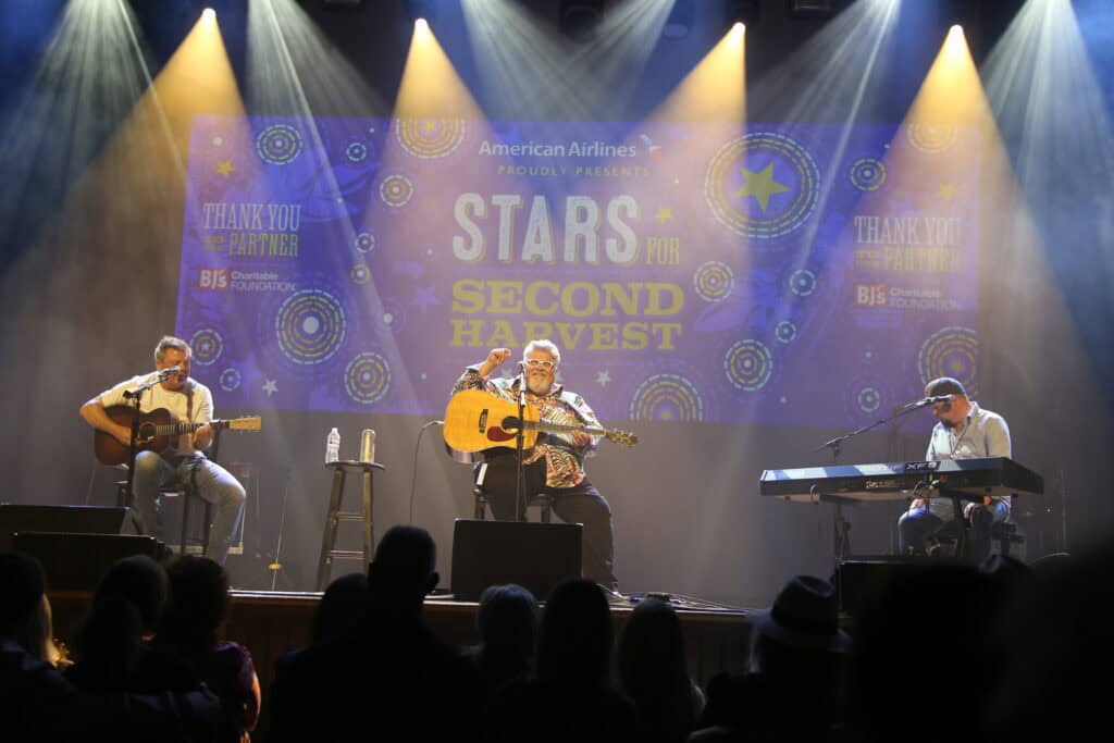 Three musicians perform Nashville Country Music on stage with guitars and a keyboard at an event called “Stars for Second Harvest,” as projected on the screen behind them, while an audience watches in the foreground.