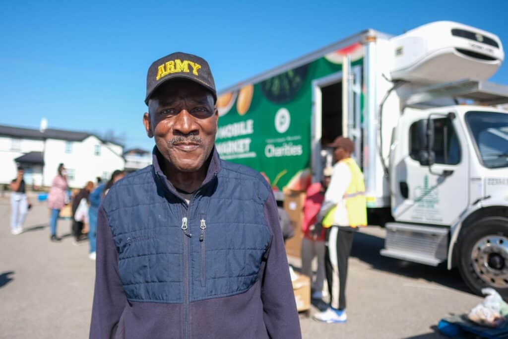 A man wearing an ARMY cap and navy vest smiles at an outdoor event, with a food bank truck and volunteers distributing boxes in the background on a sunny day.