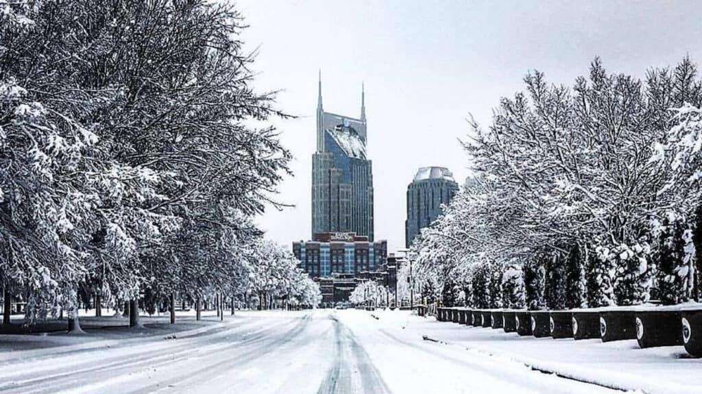 A snow-covered city street lined with trees leads toward tall buildings in the distance. Snow blankets the road, trees, and rooftops, creating a serene winter scene as a Winter Storm Threat hits Nashville under a cloudy sky.