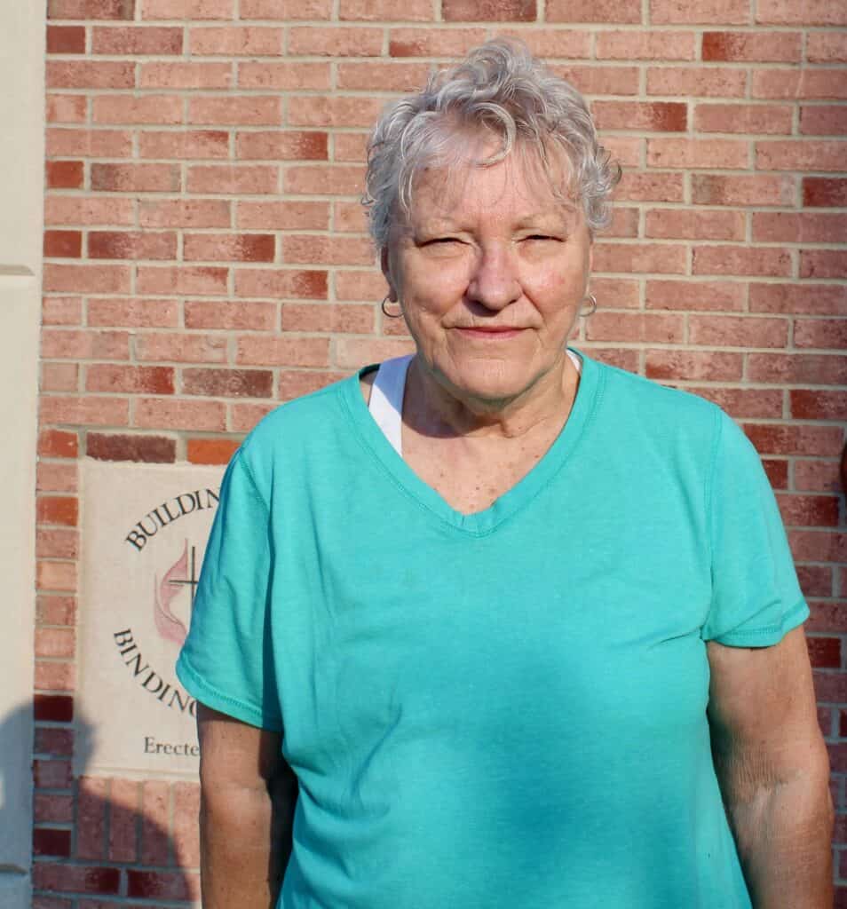 An older woman with short, curly gray hair wearing a teal shirt stands in sunlight in front of a red brick wall, with part of a building plaque visible in the background.