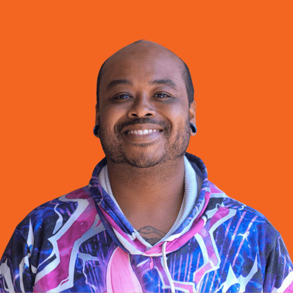 A smiling person with a short beard and stretched earlobes, wearing a colorful patterned hoodie, stands against an orange background.