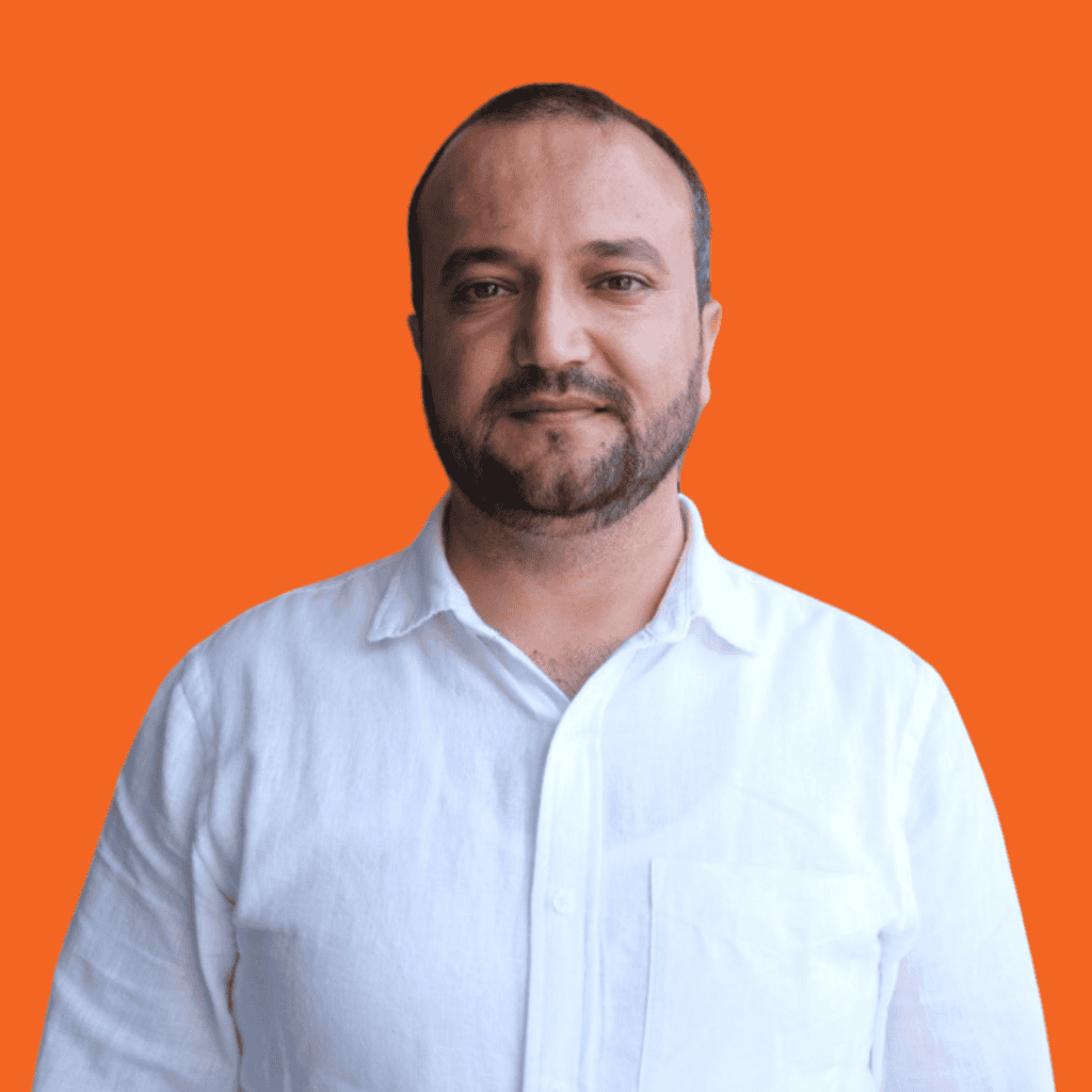 A man with short hair and a beard is wearing a white button-up shirt, standing in front of a solid orange background.