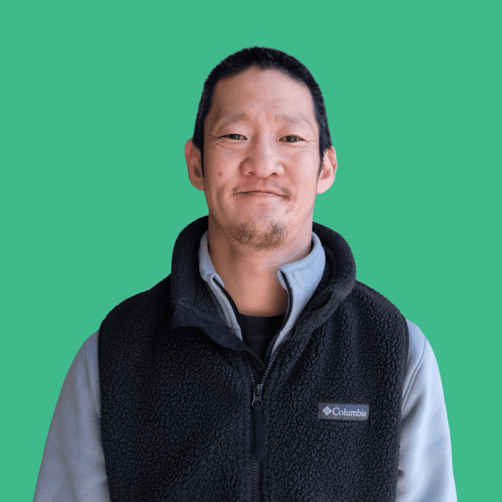 A man with short dark hair and a goatee smiles softly, wearing a gray zip-up jacket with a black Columbia fleece vest. The background is solid teal.