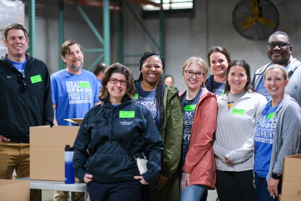 Group of volunteers smiling inside a food bank warehouse while standing beside boxes prepared for distribution.
