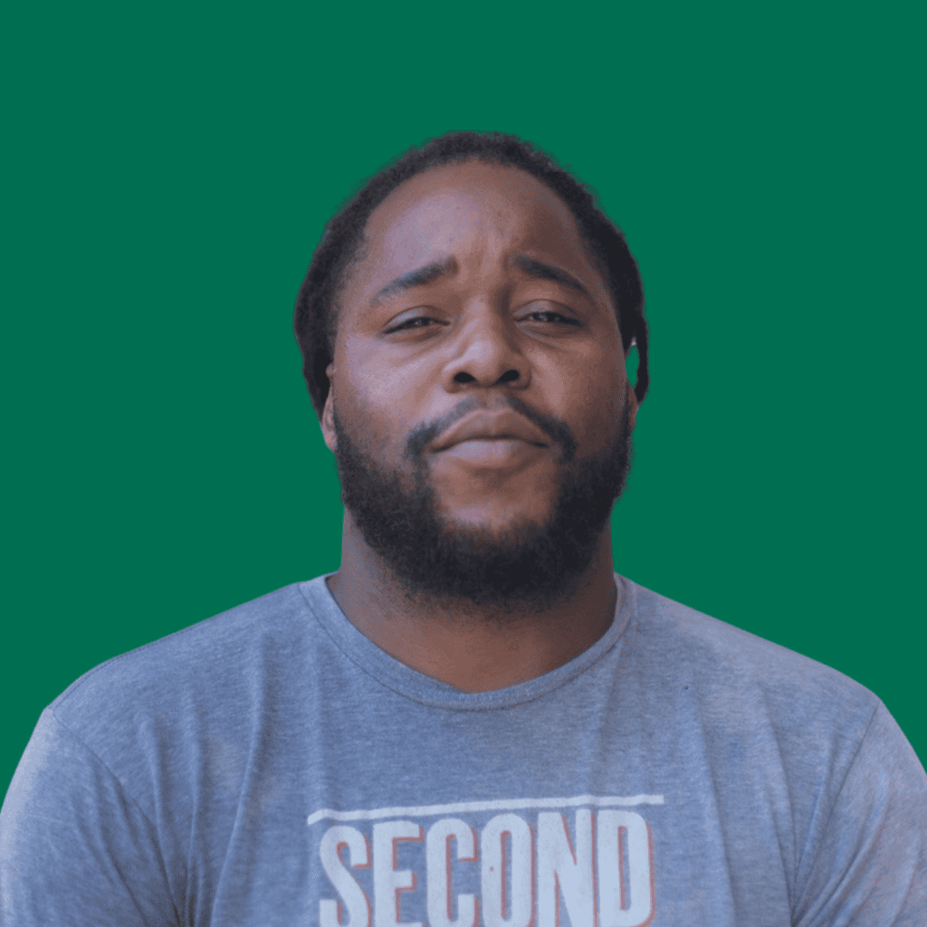 A man with a beard and mustache, wearing a gray t-shirt and looking straight ahead, stands against a solid green background.