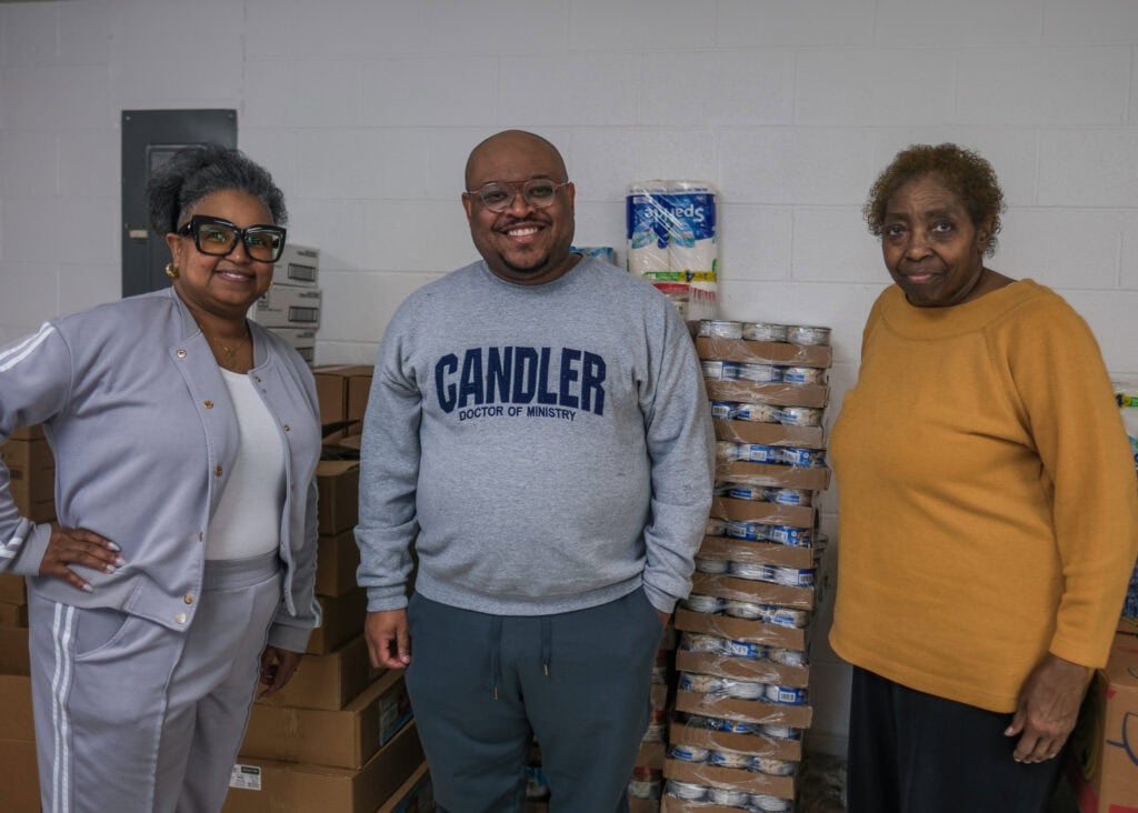 Three adults stand smiling in front of stacked canned goods and boxes, celebrating Black History in Nashville. Two women flank a man wearing glasses and a Candler Doctor of Ministry sweatshirt. The background features supplies and a white wall.