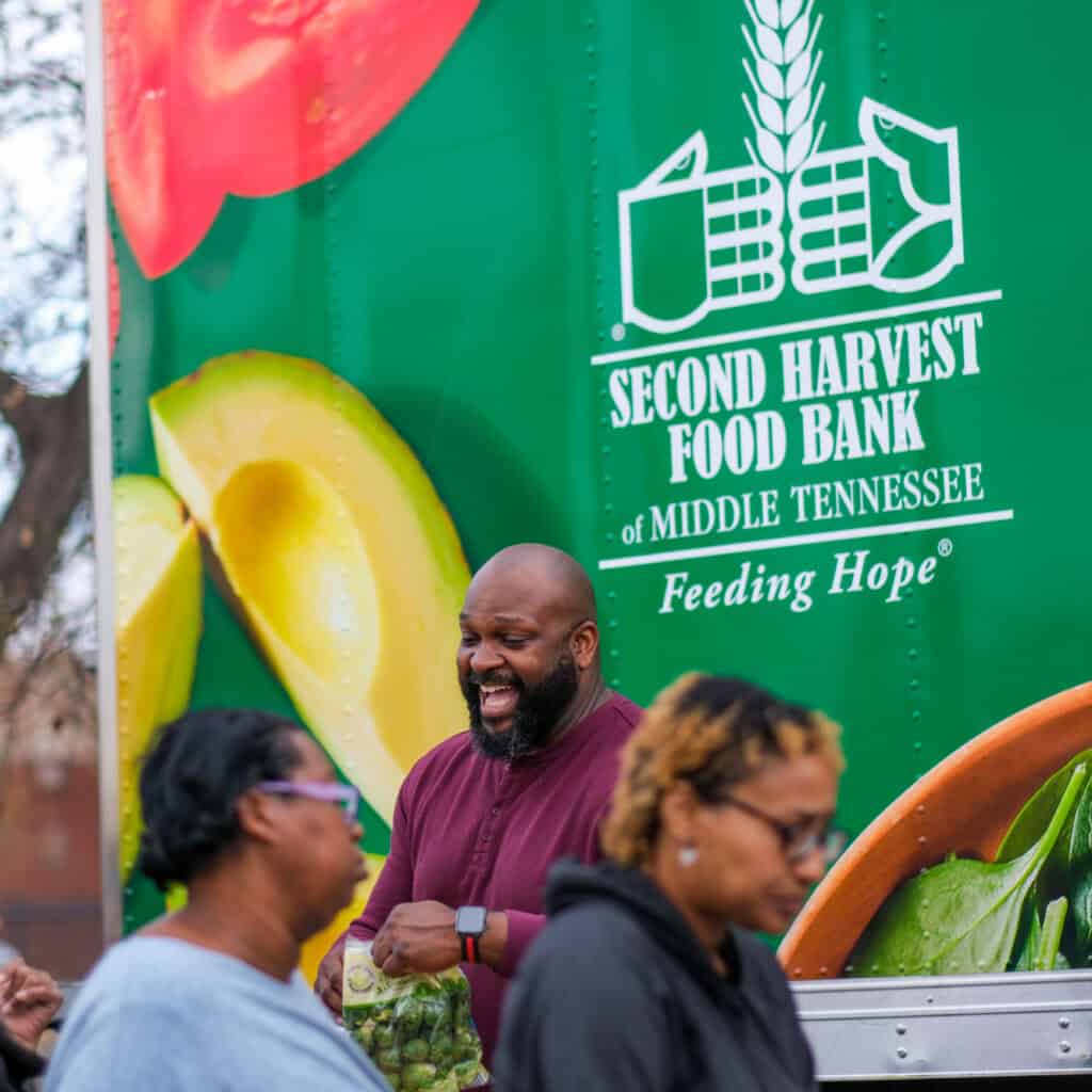 A man smiles while holding vegetables in front of a Second Harvest Food Bank of Middle Tennessee truck. Two women walk by in the foreground. Large food images decorate the truck.