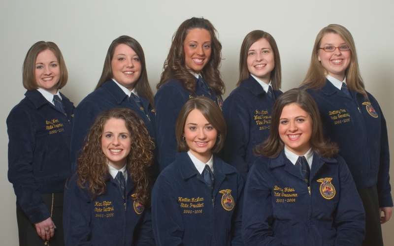 Eight young women in Women in Agriculture Leadership pose together, smiling, wearing matching official blue FFA jackets with embroidered names and ties, standing against a plain light background.