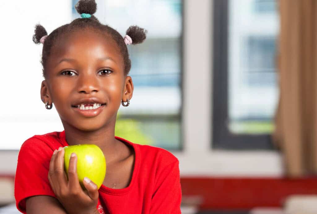 A young girl with braided hair and earrings, wearing a red shirt, smiles while holding a green apple indoors, with a blurred background of windows and curtains.