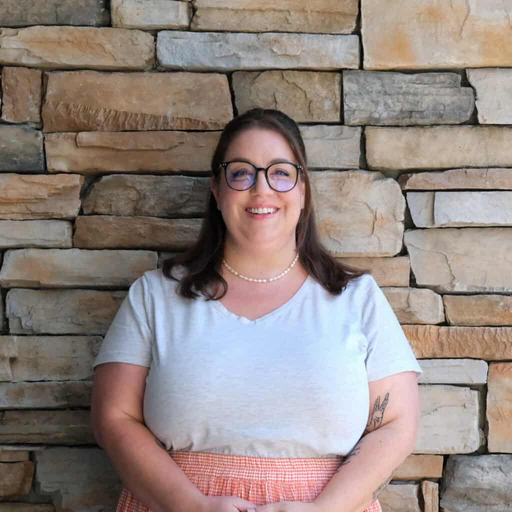 A woman with long brown hair, glasses, and a pearl necklace smiles in front of a stone wall. She is wearing a light gray T-shirt and a pink plaid skirt, with her hands clasped in front of her.