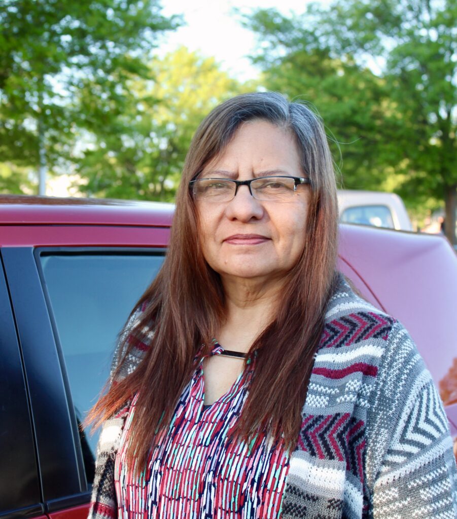 A woman with long brown hair and glasses stands outside next to a red car, wearing a patterned sweater and a striped shirt. Trees and another car are visible in the background on a sunny day.