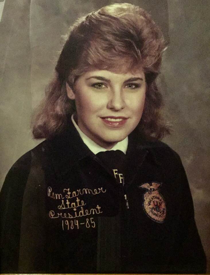 A young woman with shoulder-length blonde hair wears a dark jacket embroidered with Kim Farmer State President 1984-85 and an FFA emblem, representing Women in Agriculture Leadership. She is smiling and posing for a formal portrait with a neutral background.