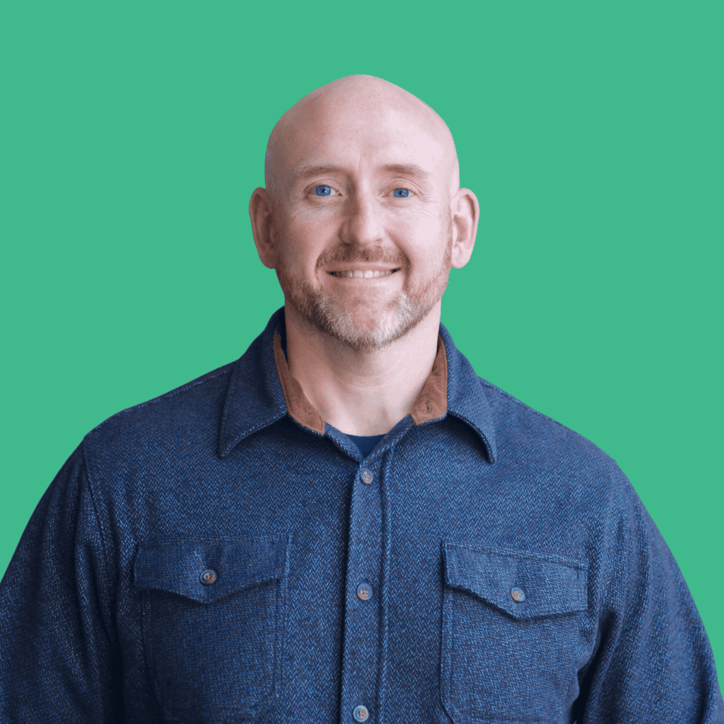 A smiling bald man with a short beard wearing a blue button-up shirt stands in front of a solid green background.