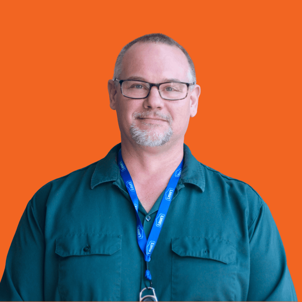 A middle-aged man with glasses, short hair, and a goatee wears a dark green shirt and a blue lanyard, standing against a solid orange background.