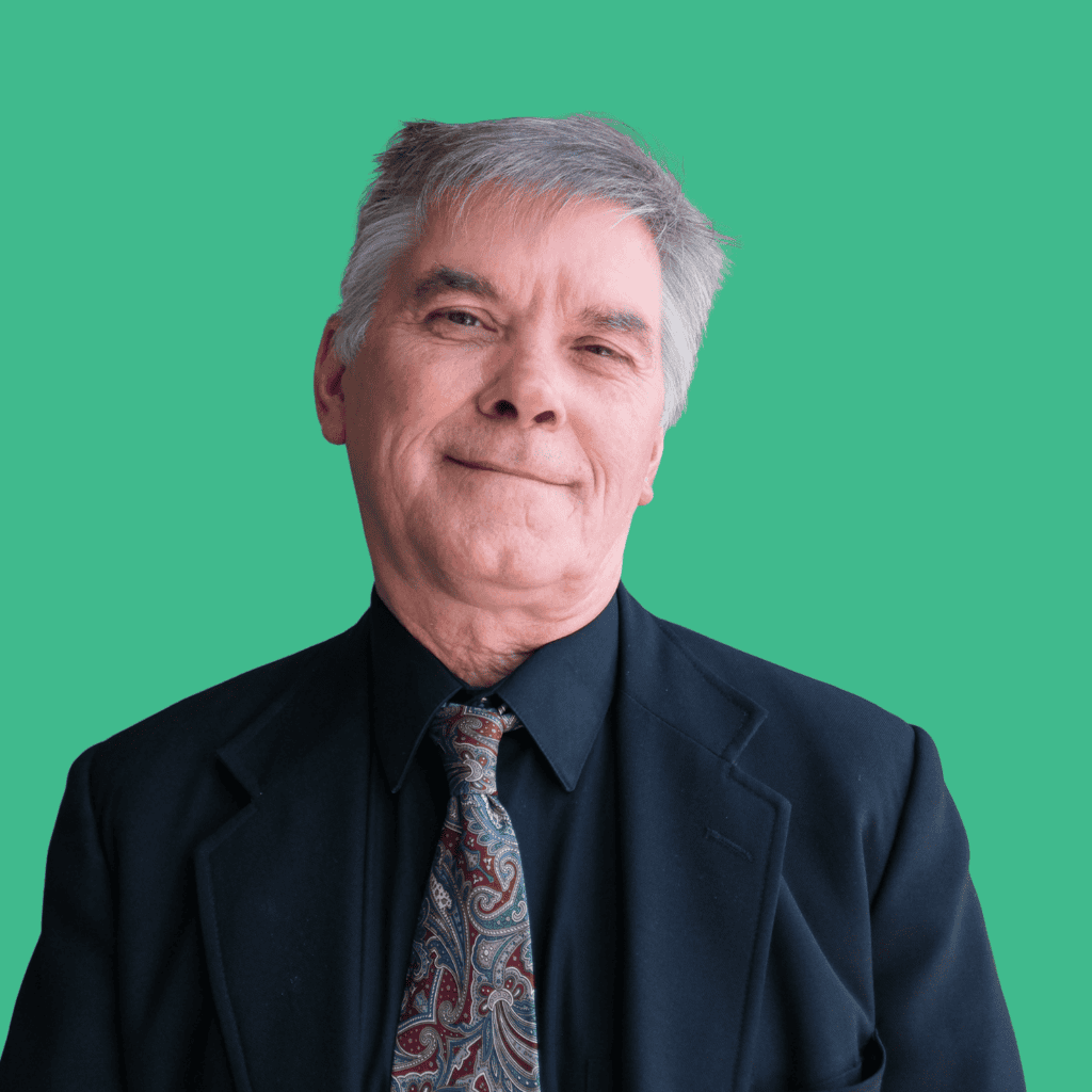 An older man with gray hair wearing a black suit, black shirt, and patterned tie stands in front of a solid teal background, smiling gently at the camera.