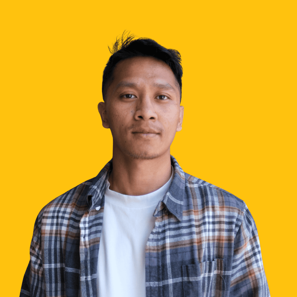 A person with short dark hair wearing a plaid shirt over a white t-shirt stands against a solid yellow background, looking directly at the camera with a neutral expression.