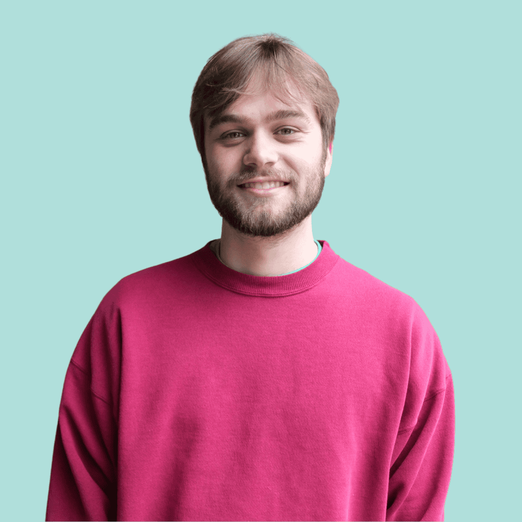 A young man with light brown hair and a beard smiles while wearing a red sweatshirt, standing in front of a solid light blue background.