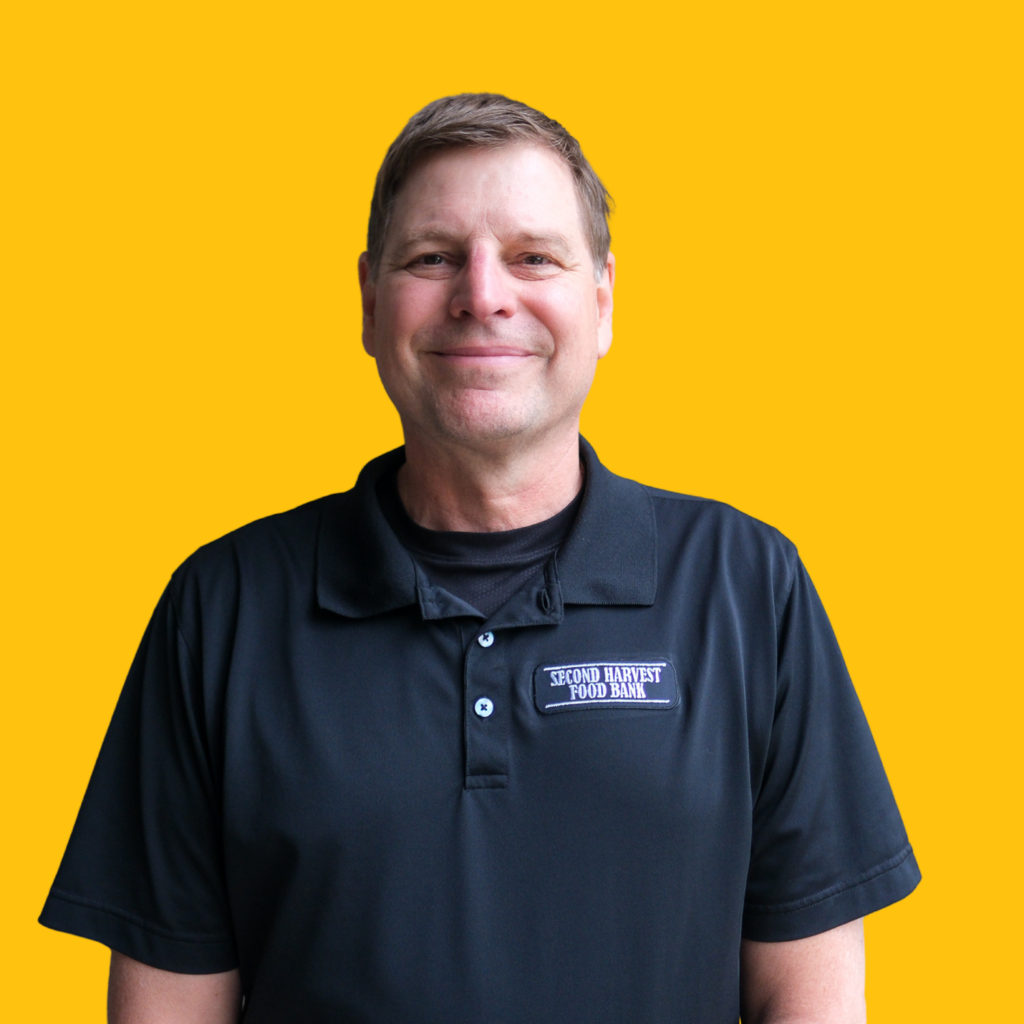 A man wearing a black Second Harvest Food Bank polo shirt stands smiling in front of a solid yellow background.