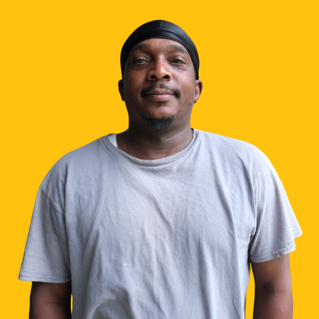 A man wearing a light gray T-shirt and a black headscarf stands in front of a solid yellow background, looking at the camera with a neutral expression.