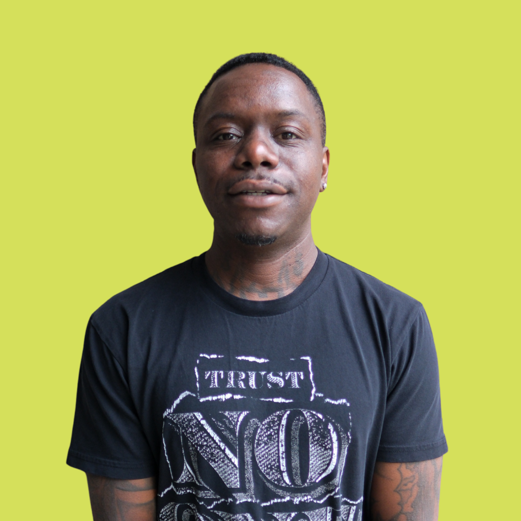 A person with short hair wearing a black t-shirt with white text stands against a plain light green background, facing the camera and smiling slightly.