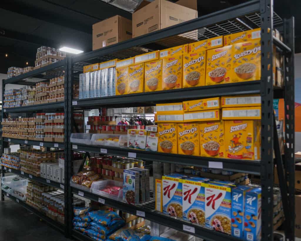 Metal shelves in a store stocked with various food items, including boxes of Toastys and Scooters cereal, crackers, bags of snacks, canned goods, and other packaged products.