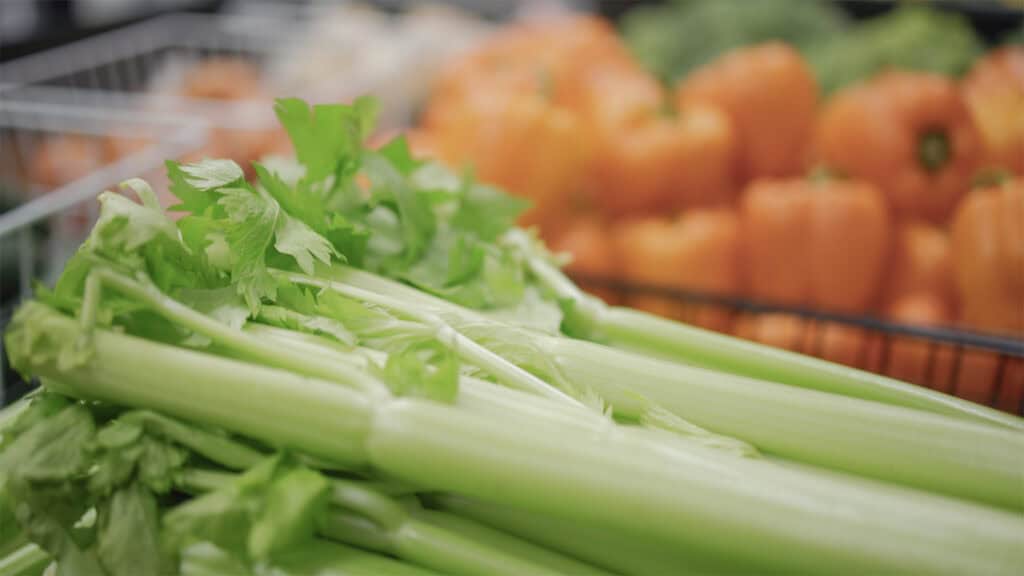 Fresh celery stalks with leafy tops are displayed in the foreground, while orange bell peppers are visible, slightly out of focus, in the background at a grocery store or market.