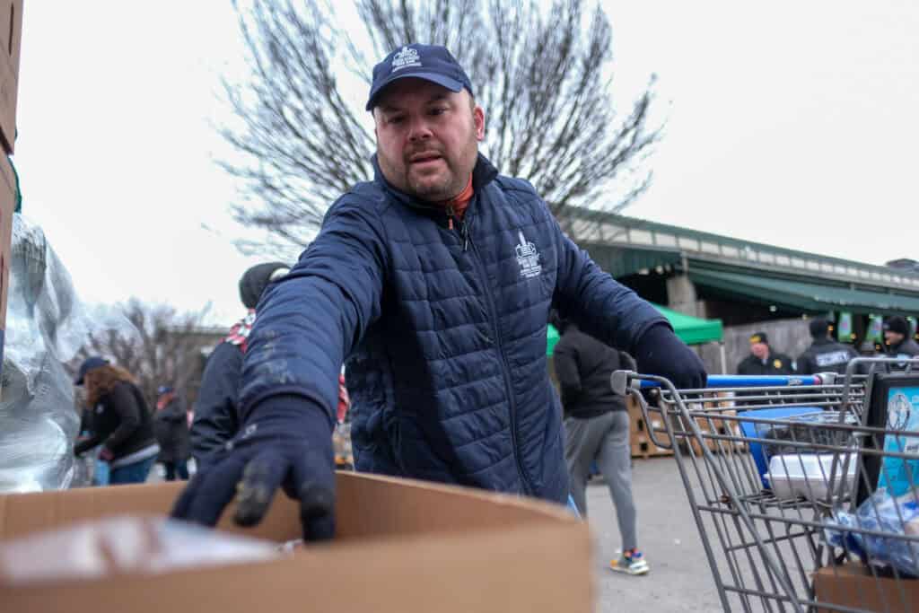 A man wearing a navy jacket, hat, and gloves places items into a shopping cart outdoors, with other people and trees visible in the background.