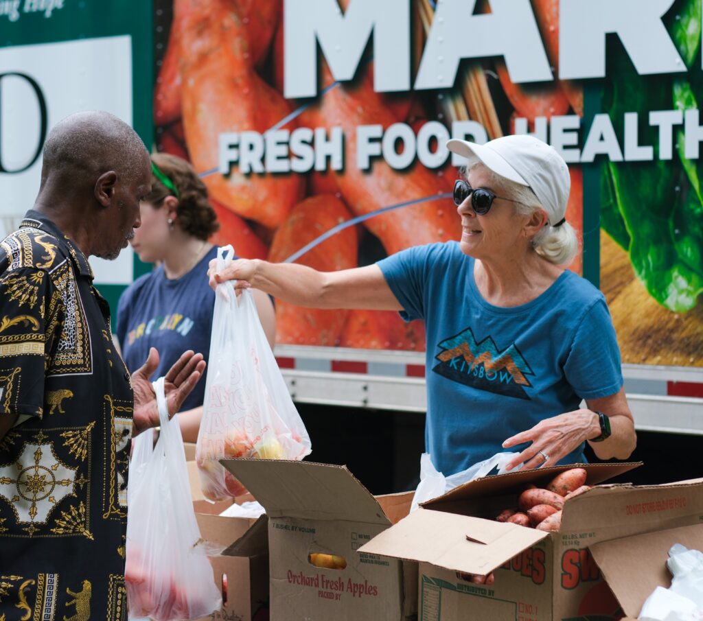 A smiling woman wearing sunglasses hands a bag of produce to a man at an outdoor food distribution event, with boxes of fruits and vegetables on the table and a large “Fresh Food” sign in the background.