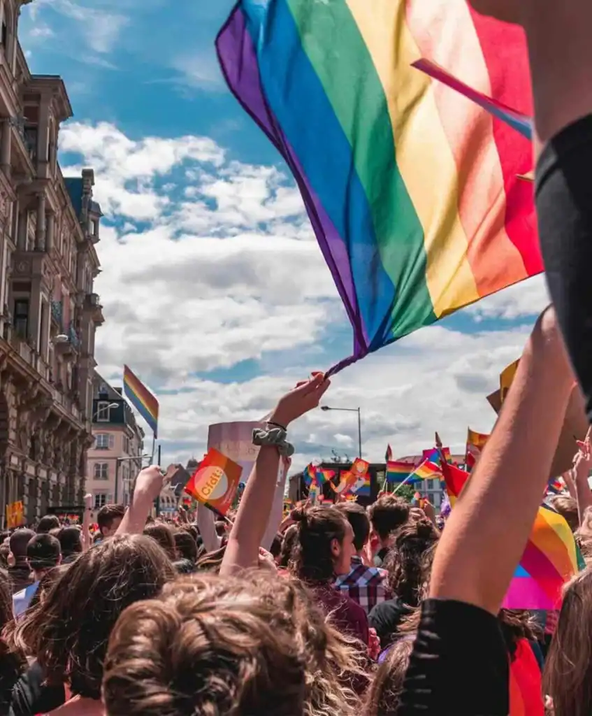A crowd of people at a pride parade wave rainbow flags under a blue sky with clouds, surrounded by historic buildings and holding colorful signs and banners.