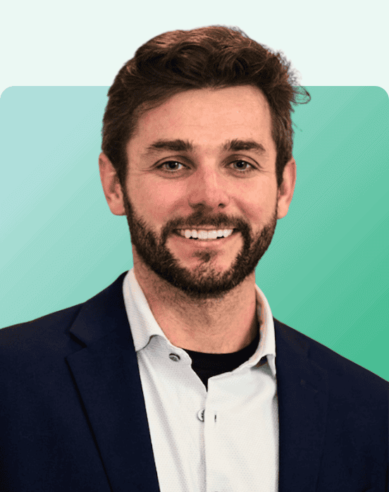 A man with short brown hair and a beard, wearing a navy blazer over a light shirt, smiling in front of a teal gradient background.