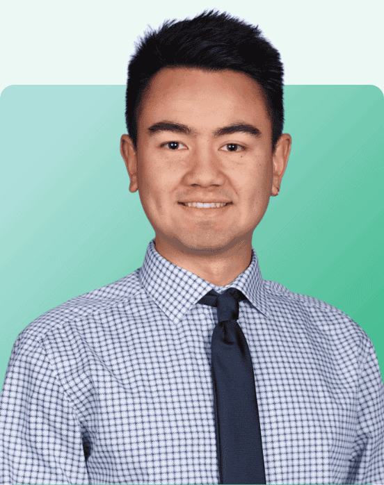 A man with short dark hair, wearing a checkered white and blue shirt and a dark tie, smiles at the camera against a teal gradient background.