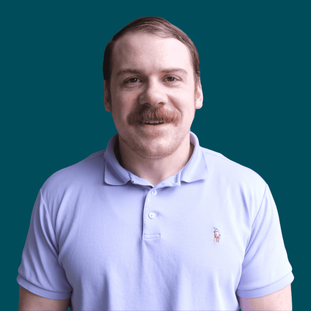 A smiling man with short hair and a mustache, wearing a light purple polo shirt, stands in front of a teal background.