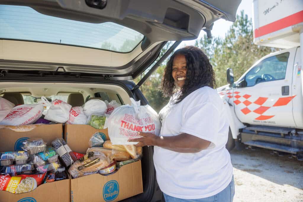 A woman smiles as she loads groceries and packaged food from a car trunk. A moving truck is parked nearby. She wears a white shirt and stands outdoors on a sunny day.