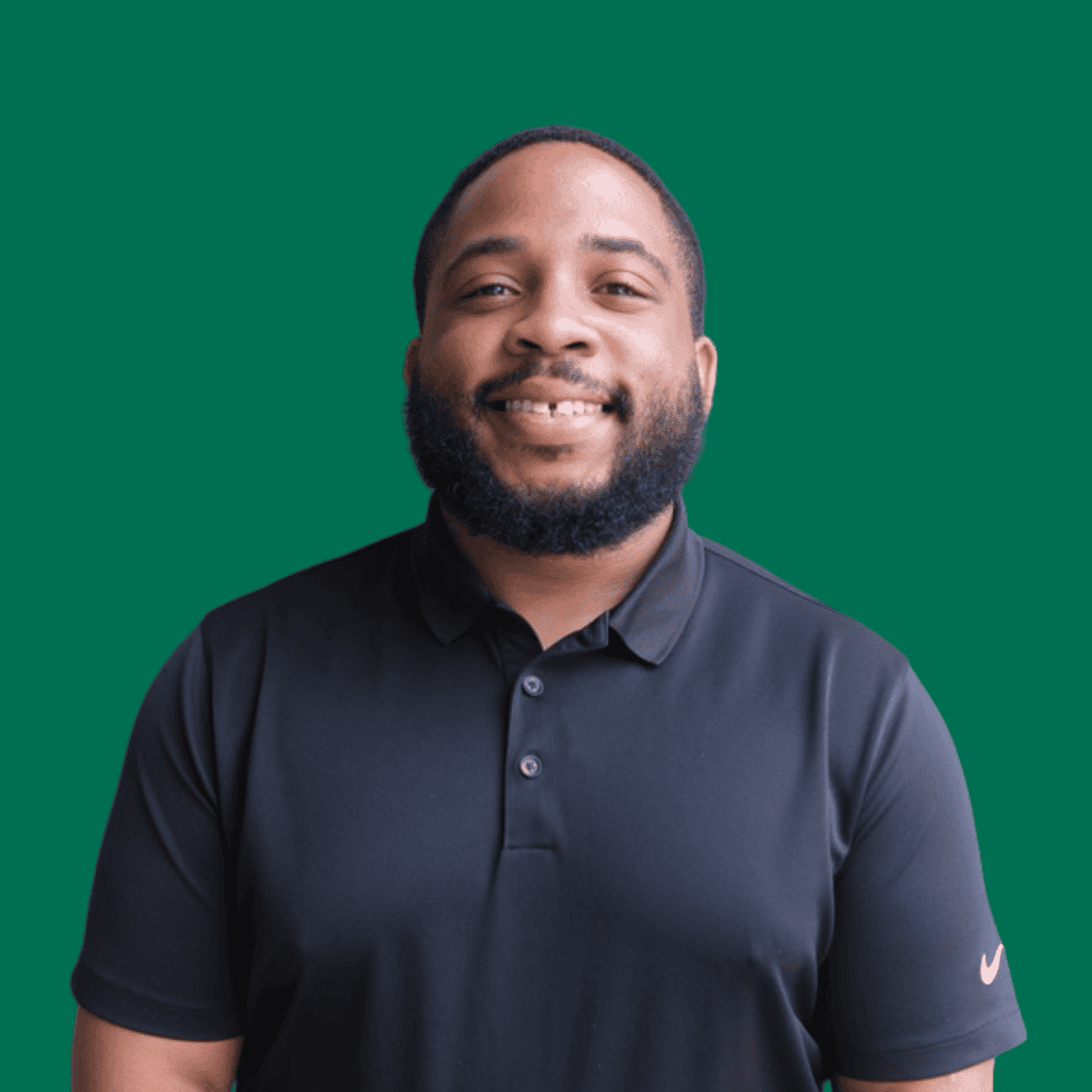 A man with a beard and mustache smiles, wearing a black Nike polo shirt, standing in front of a solid green background.