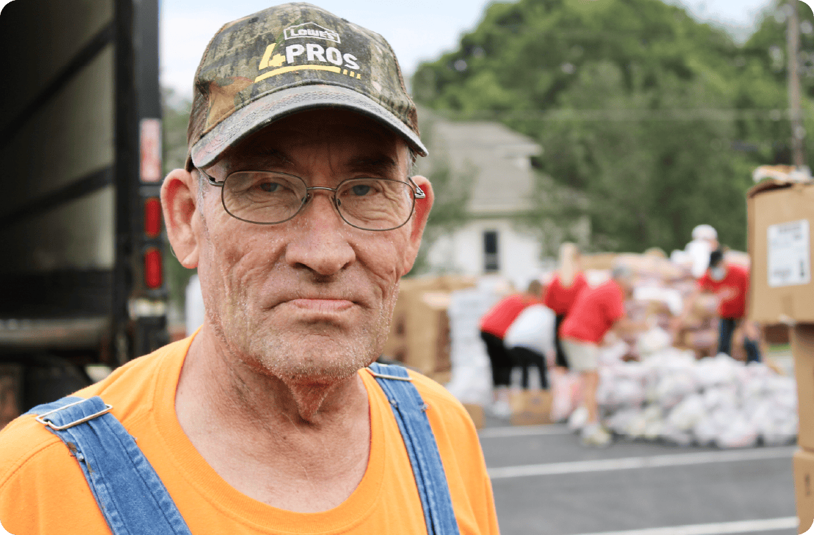 An older man in glasses, an orange shirt, and a camouflage hat stands in front of a group sorting supplies outdoors near a truck.