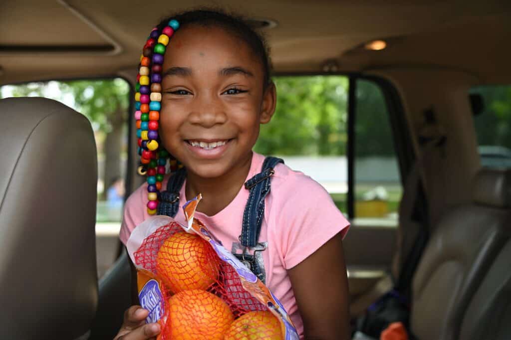 A smiling girl with colorful beads in her hair sits in a car holding a bag of oranges.