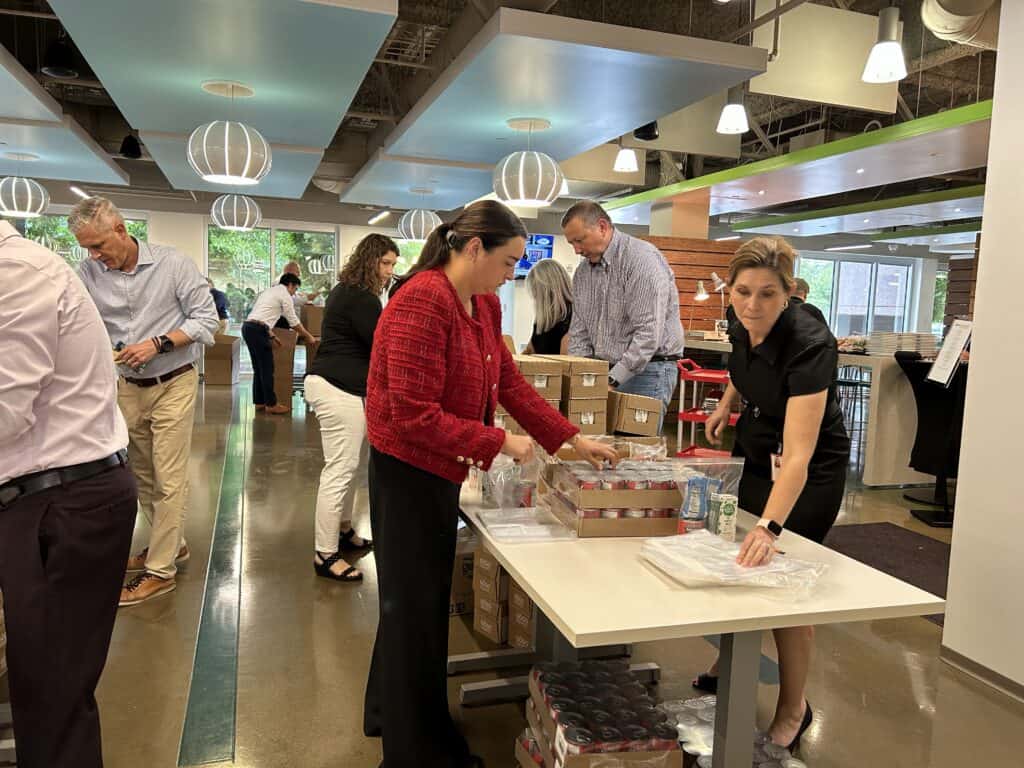 People in business attire pack food and supplies into bags at tables in a bright, modern office space, participating in a group volunteer or charity event.