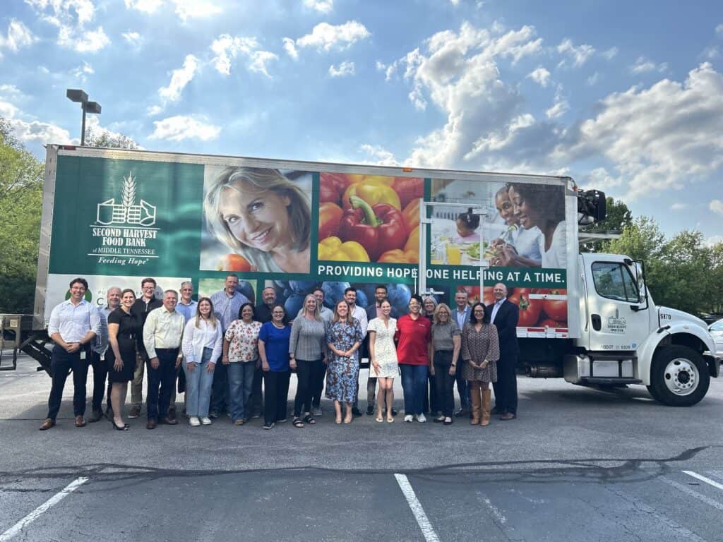 A group of people stands smiling in front of a Second Harvest Food Bank truck with colorful produce and happy faces displayed on its side, in a parking lot under a partly cloudy sky.