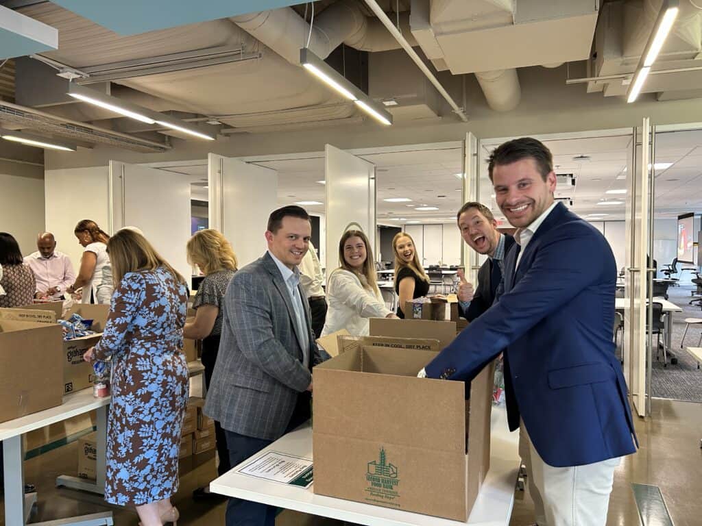 A group of professionally dressed people smiling as they pack items into cardboard boxes in a modern office setting.