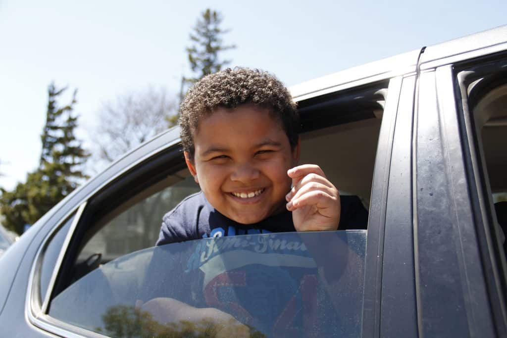 A smiling child leans out of a car window on a sunny day, holding up their fingers as if waving. Trees and a clear blue sky are visible in the background.