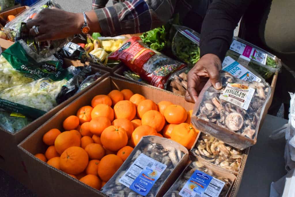 Two people pick out groceries from boxes filled with oranges, salad greens, packaged mushrooms, and other produce at an outdoor market or food distribution event.